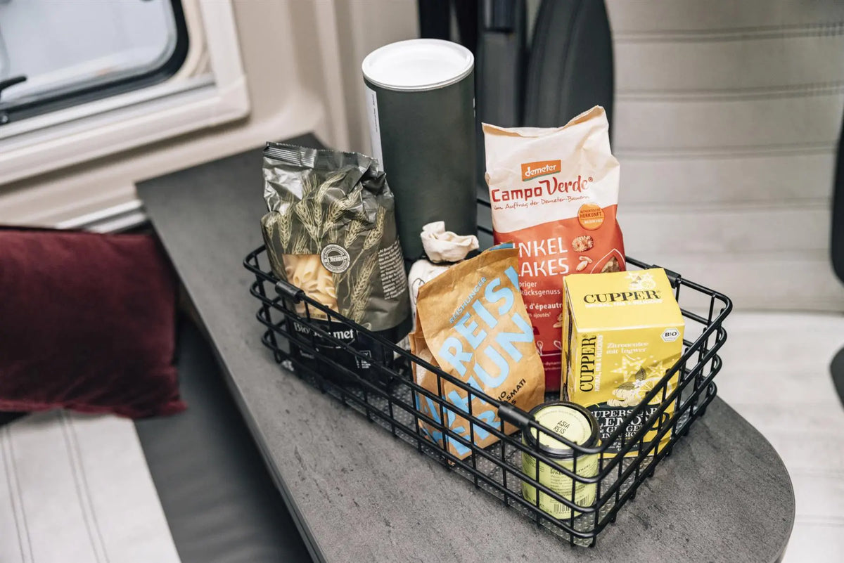 Black wire Sunlight motorhome basket with snacks and a cup on a vehicle's dashboard.